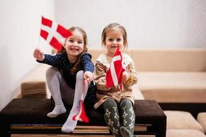 duas irmãs estão sentadas em um sofá em casa com bandeiras dinamarquesas nas mãos. Dinamarca crianças meninas com bandeira. foto