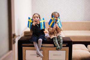 duas irmãs estão sentadas em um sofá em casa com bandeiras suecas nas mãos. Suécia crianças meninas com bandeira. foto