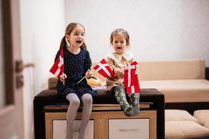 duas irmãs estão sentadas em um sofá em casa com bandeiras dinamarquesas nas mãos. Dinamarca crianças meninas com bandeira. foto