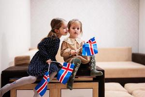 duas irmãs estão sentadas em um sofá em casa com bandeiras islandesas nas mãos. Islândia crianças meninas com bandeira. foto