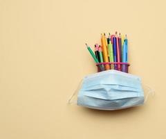 máscara médica descartável e lápis e canetas multicoloridos em um balde de metal em um fundo bege foto