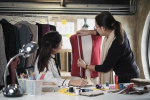designer de moda feminina asiática de meia-idade ensina alfaiate jovem estagiário adolescente sobre forma e tamanho com fita métrica e tecido de costura para coleção de design de vestido, pequena empresa boutique profissional. foto