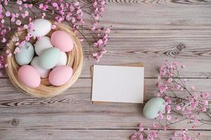 flores cor de rosa, um ninho com ovos em tons pastel suaves e cartão branco em branco para texto em um fundo de madeira. brincar. lugar para texto. cartão de páscoa foto