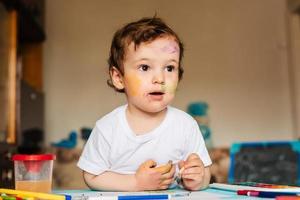 um menino bonito pinta com pincéis e tintas coloridas em uma folha de papel foto