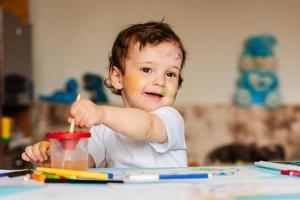 um menino bonitinho desenha com pincéis e tintas coloridas em uma folha de papel foto