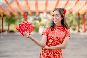 feliz Ano Novo Chinês. mulher asiática usando vestido cheongsam qipao tradicional parecendo confiante segurando ang pao, envelopes vermelhos no templo budista chinês. texto chinês significa muita sorte grande lucro foto