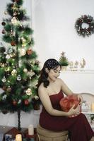 retrato de uma jovem bonita, confortável, sente-se segurando o presente de natal, sorrindo, use um vestido vermelho na sala de estar decorada de natal dentro de casa foto