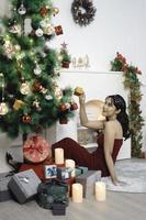 retrato de uma jovem bonita, confortável, sente-se segurando o presente de natal, sorrindo, use um vestido vermelho na sala de estar decorada de natal dentro de casa foto