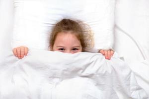 menina com olhos sorridentes na cama escondida debaixo das cobertas foto