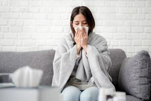 mulher asiática doente coberta com cobertor espirrando sentada no sofá ela estava doente e com febre. conceito de gripe foto