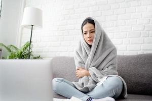 jovem chateado congelando, sentindo frio em casa, sentado no sofá coberto com cobertor foto