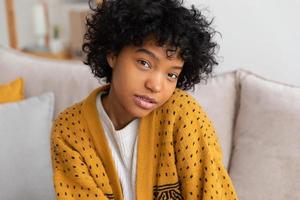linda garota afro-americana com penteado afro sorrindo sentado no sofá em casa interior. jovem africana com cabelos cacheados rindo. liberdade felicidade despreocupado conceito de pessoas felizes.