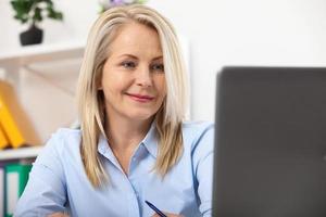 trabalhando em novo projeto. mulher de negócios trabalhando no escritório com documentos. linda mulher de meia idade olhando para laptop com sorriso enquanto está sentada no escritório. foto