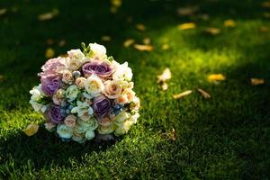 foto de um buquê de casamento, buquê de rosas brancas e rosas, flores brancas e amarelas com verde, decoradas com fitas de seda, deitam-se na grama verde. o buquê da noiva.