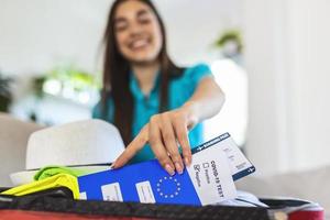 mulher feliz planejando uma viagem durante a pandemia de covid-19, preparando uma mala. mulher preparando a viagem de férias. segurando passaporte, passagem e teste negativo de coronavírus na mão foto