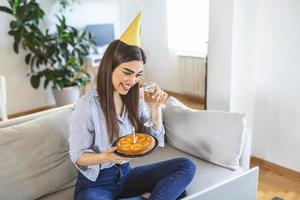 novo conceito normal. mulher feliz comemorando seu aniversário sozinha. jovem mulher segurando um bolo. festa de aniversário em casa. chamada de vídeo em família. distanciamento social. vida em casa. foto