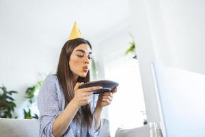 novo conceito normal. mulher feliz comemorando seu aniversário sozinha. jovem mulher segurando um bolo. festa de aniversário em casa. chamada de vídeo em família. distanciamento social. vida em casa. fazendo um desejo