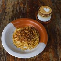 prato saudável de espaguete italiano coberto com um saboroso tomate e molho à bolonhesa de carne moída e manjericão fresco em uma mesa de madeira marrom rústica. servido com capuccino foto