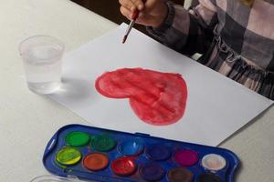 menina sentada à mesa e desenhando aquarela de coração vermelho, cartão postal de dia dos namorados, cartão de felicitações, conceito de escola de arte, publicidade, criança pintando aquarela em casa, atividade infantil, lazer infantil. foto