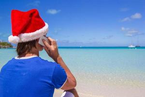 jovem de chapéu de Papai Noel falando no telefone na praia tropical foto