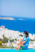 mulher magra aplicando protetor solar nas pernas, sentada à beira da piscina, fundo da cidade velha de mykonos, na europa foto