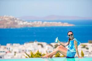 jovem aplicando protetor solar nas pernas, sentada à beira da piscina, fundo da velha cidade de mykonos, na europa foto
