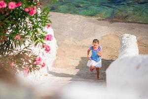 adorável garota se divertindo ao ar livre. garoto na rua da típica vila tradicional grega com paredes brancas e portas coloridas na ilha de mykonos, na grécia foto