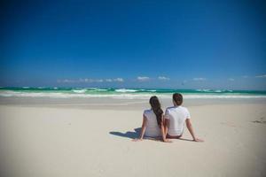 vista traseira do jovem casal sentado na praia tropical branca foto