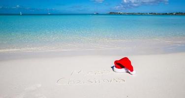 chapéu de papai noel na praia de areia branca e feliz natal escrito na areia foto