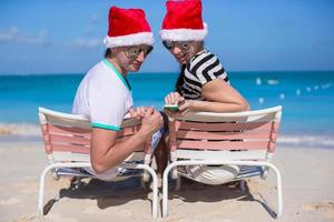 família de dois em chapéus de papai noel sentado na cadeira de praia foto