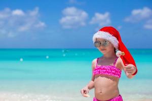 menina bonitinha com chapéu de Papai Noel vermelho na praia tropical foto