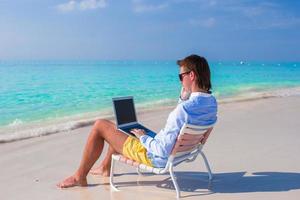 jovem com laptop e celular na praia tropical foto