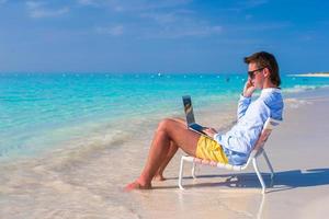 jovem com laptop e celular na praia tropical foto