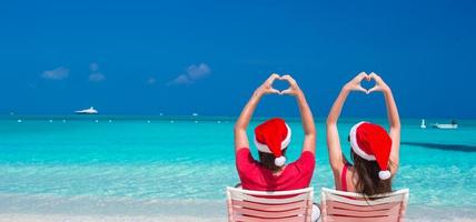 casal romântico feliz em chapéus de papai noel vermelhos na praia fazendo corações foto