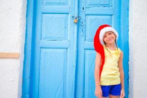 menina de chapéu de papai noel ao ar livre na velha rua nas férias de natal em mykonos. garoto na rua da típica vila tradicional grega com paredes brancas e portas coloridas na ilha de mykonos, na grécia foto