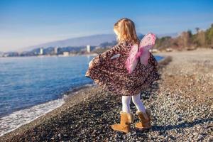 menina adorável com asas de borboleta correndo ao longo da praia em um dia ensolarado de inverno foto