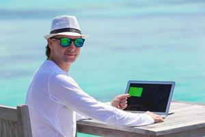 jovem trabalhando no laptop com cartão de crédito na praia tropical foto