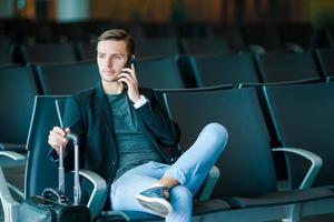 homem de negócios urbano falando no telefone inteligente viajando dentro do aeroporto. casual jovem empresário vestindo paletó. belo modelo masculino. jovem com celular no aeroporto enquanto aguarda o embarque. foto