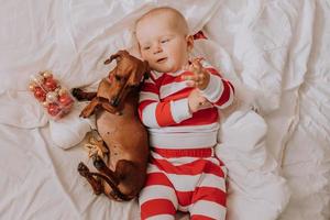 menino de pijama vermelho e branco está deitado na cama em um lençol branco com um cachorro. manha de Natal. sono saudável da criança. espaço para texto. foto de alta qualidade