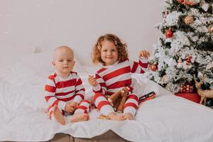 crianças de pijama vermelho e branco comem doces de natal sentados na cama. irmão e irmã, menino e menina compartilham presentes. manha de Natal. estilo de vida. espaço para texto. foto de alta qualidade