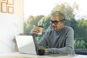 retrato idoso homem asiático sênior senta-se perto da janela de vidro pela manhã para trabalhar em casa e verificando seus negócios em seu laptop na mesa com foco sério, suave e seletivo. foto