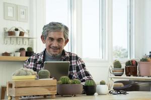 retrato idoso homem asiático sênior senta-se perto da janela de vidro pela manhã para trabalhar em casa e verificando seus negócios em seu laptop na mesa com foco sério, suave e seletivo. foto