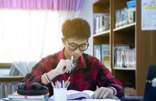 retrato jovem rapaz asiático usando óculos e pulseira de arco-íris, segurando a caneta, sentado na biblioteca, lendo livros e concentrando-se para reservar na mesa antes do teste intermediário e teste final no dia seguinte. foto