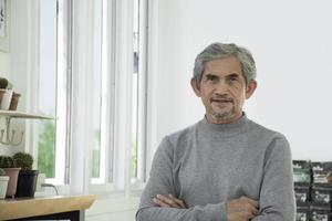 retrato idoso homem asiático sênior senta-se perto da janela de vidro pela manhã para trabalhar em casa e verificando seus negócios em seu laptop na mesa com foco sério, suave e seletivo. foto