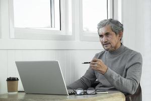 retrato idoso homem asiático sênior senta-se perto da janela de vidro pela manhã para trabalhar em casa e verificando seus negócios em seu laptop na mesa com foco sério, suave e seletivo. foto