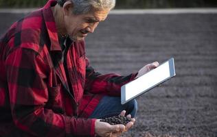 agricultor de grãos de café inteligente asiático idoso em camisa vermelha e preta, sentado na área do meio do chão de secagem de pilha de grãos de café e segurando o taplet na mão para verificar a qualidade do café, foco suave e seletivo. foto