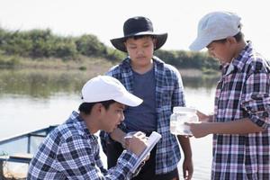 jovem garoto asiático segura tubo transparente que tem água de exemplo dentro para fazer o experimento e medição do nível de ph enquanto seu projeto escolar trabalha com seus amigos atrás no rio onde ele morava. foto