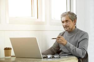 retrato idoso homem asiático sênior senta-se perto da janela de vidro pela manhã para trabalhar em casa e verificando seus negócios em seu laptop na mesa com foco sério, suave e seletivo. foto