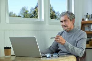 retrato idoso homem asiático sênior senta-se perto da janela de vidro pela manhã para trabalhar em casa e verificando seus negócios em seu laptop na mesa com foco sério, suave e seletivo. foto