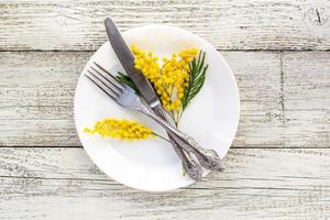 prato de configuração de mesa festiva com garfo e faca e decoração de flores mimosa em fundo branco de madeira com espaço de cópia foto